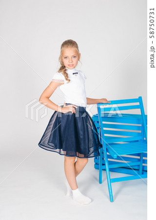 Cute girl with long blond curly hair in school fashion clothes with blue chair on a white background Cute girl with long blond curly hair in school fashion clothes with blue chair on a white background 58750731