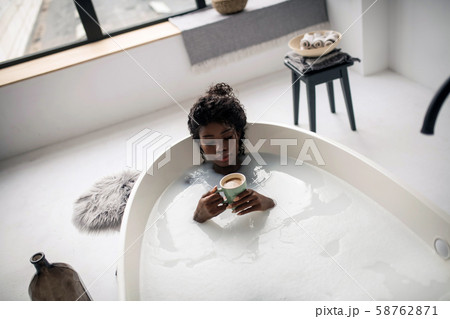 Curly dark-skinned woman drinking cappuccino while lying in bath Curly dark-skinned woman drinking cappuccino while lying in bath 58762871