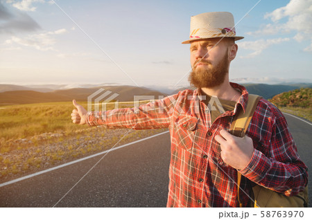 Stylish barefoot bearded male hitchhiker traveler in a hat and with a backpack walks along a 58763970
