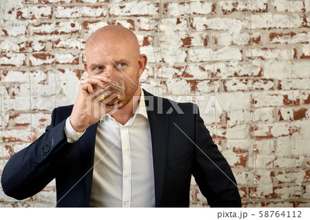 A man in a business suit on a background of a brick wall drinks a drink from a glass. The concept of 58764112