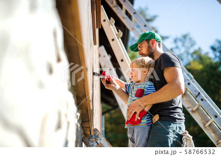 A father and toddler boy outdoors in summer, painting wooden house. A father and toddler boy outdoors in summer, painting wooden house. 58766532
