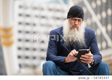 Mature bearded businessman sitting while using phone in the city outdoors Mature bearded businessman sitting while using phone in the city outdoors 58785447