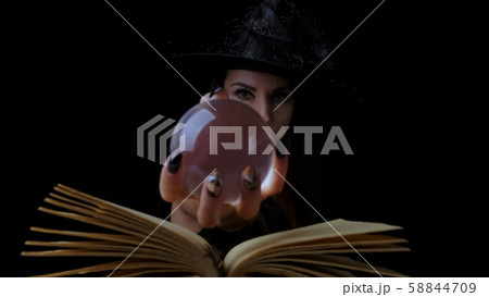 female in a black witch costume and hat, holds in her hands a transparent magic ball over a book and female in a black witch costume and hat, holds in her hands a transparent magic ball over a book and 58844709