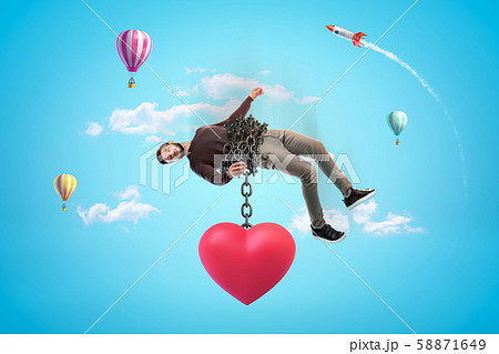 Man in blue sky bound with chains to huge red heart which is pulling him down. 58871649