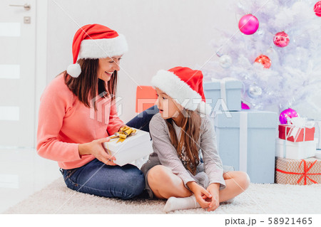 Mother in santa hat is going to give her lovely daughter a present, daughter is looking at the box Mother in santa hat is going to give her lovely daughter a present, daughter is looking at the box 58921465