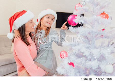 Mother holds her daughter, standing near christmas tree, cute girl is pointing at one of pink 58921466