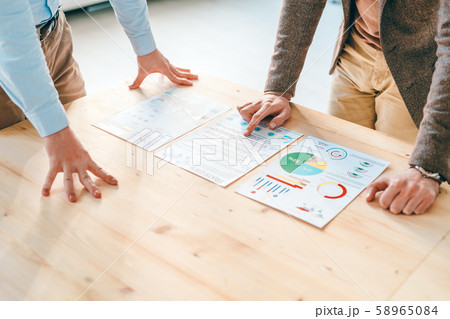 Two young economists in formalwear bending over financial papers on wooden table Two young economists in formalwear bending over financial papers on wooden table 58965084