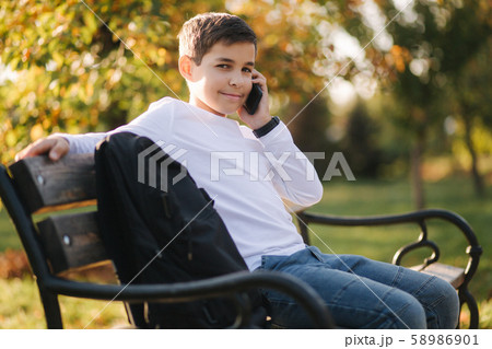 Young boy in white sweatshirt with black backpack sitting on the bench in the park and speak with Young boy in white sweatshirt with black backpack sitting on the bench in the park and speak with 58986901