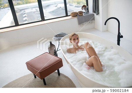 Smiling woman enjoying foamy bath standing near window 59002184