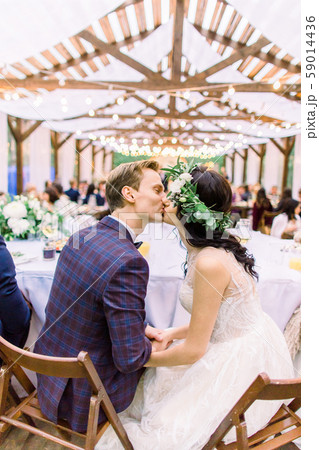Bride and groom sitting at the table and kissing, with guests at wedding reception outside in the 59014436