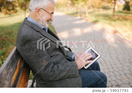 Handsome gray-haired elderly man with beautiful beard sitting on the bench and use tablet for 59037791