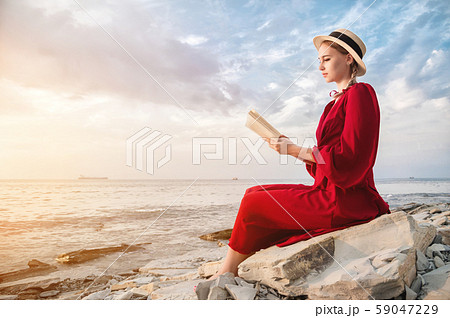 An attractive Caucasian girl in a red bright dress and a straw hat sits on a large stone by the sea 59047229