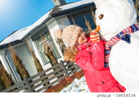 Winter vacation. Girl standing outdoors near house making snowman nose with carrot smiling happy Winter vacation. Girl standing outdoors near house making snowman nose with carrot smiling happy 59058538