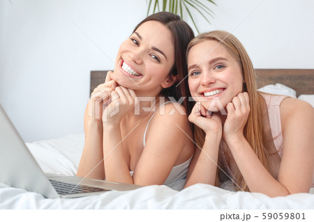 Friends together at home lying on bed with laptop posing to camera joyful Friends together at home lying on bed with laptop posing to camera joyful 59059801