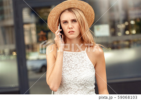 Traveler. Young woman in hat standing on street talking on smartphone looking camera serious Traveler. Young woman in hat standing on street talking on smartphone looking camera serious 59068103
