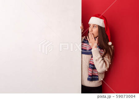 Freestyle. Young woman wearing scarf and santa hat standing isolated on red looking at white board 59073004