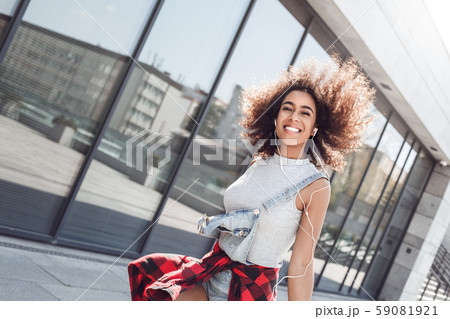 Young woman in the city street jumping near window with smartphone wearing earphones listening music 59081921