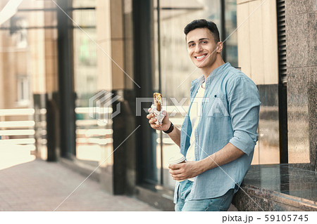 Outdoors Leisure. Stylish guy standing on the street with cup of coffee eating sandwich laughing 59105745