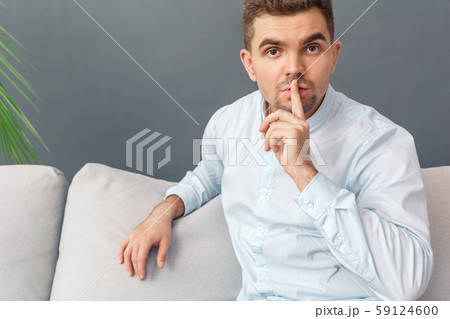 Keep Secret. Young man studio sitting on sofa isolated on grey showing silence gesture to camera Keep Secret. Young man studio sitting on sofa isolated on grey showing silence gesture to camera 59124600
