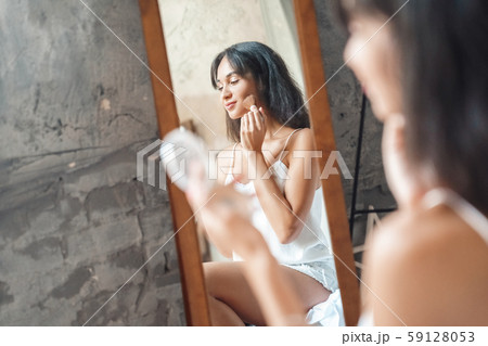 Weekend ar home. Young woman sitting on bed applying face powder smiling playful mirror reflection Weekend ar home. Young woman sitting on bed applying face powder smiling playful mirror reflection 59128053