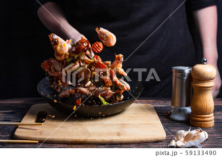 Chef at work. Hand holds a hot pan with fried shrimp and vegetables in sweet and sour sauce. Process of preparation of shrimps over dark background Chef at work. Hand holds a hot pan with fried shrimp and vegetables in sweet and sour sauce. Process of preparation of shrimps over dark background 59133490