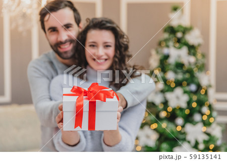 Defocused man and woman holding a present tied with a red ribbon, looking at the camera smilling. Defocused man and woman holding a present tied with a red ribbon, looking at the camera smilling. 59135141