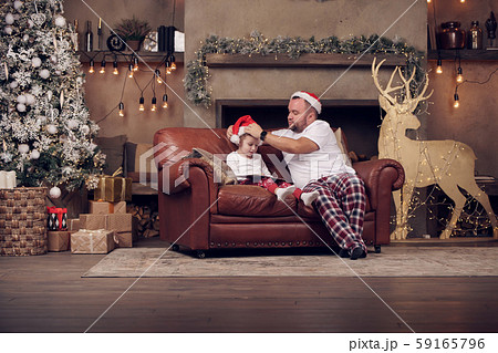 Photo of father in Santa hat and son on red sofa in room Photo of father in Santa hat and son on red sofa in room 59165796