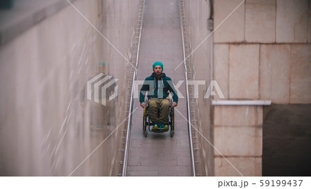 Disabled man in wheelchair wearing jacket and hat - going down the long ramp in the subway 59199437