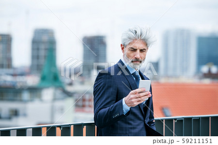 A mature businessman standing on a terrace, using smartphone. 59211151