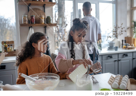 Sister pouring milk into glass while cooking pie with sister 59228499
