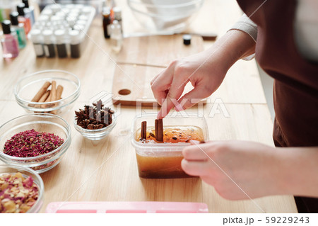 Hands of young woman putting cinnamon sticks into liquid soap mass in container 59229243