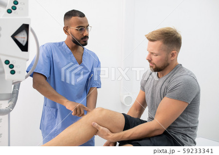 Young doctor in uniform consulting sportsman while pointing at his sick leg Young doctor in uniform consulting sportsman while pointing at his sick leg 59233341
