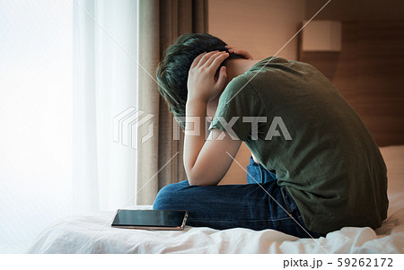 Cyberbullying concept. Young Asian preteen/teenage boy sit cross legged alone next to a smartphone on the bed, covering his ears with hands. Stressed, frustrated, overwhelmed with online bullying. 59262172