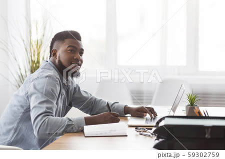 Black man taking notes from laptop at workplace in office Black man taking notes from laptop at workplace in office 59302759