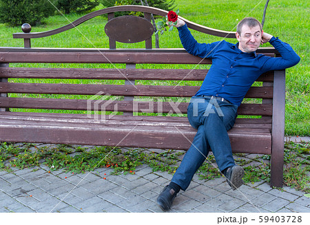 A man with a rose relaxing sitting on a park bench and winking at women passing by 59403728