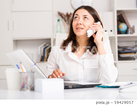 Young female using laptop and talking by smartphone at the office Young female using laptop and talking by smartphone at the office 59429057