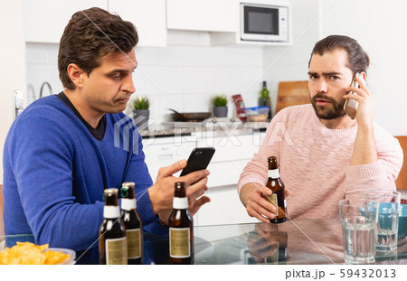 Two concerned men talking and laughing while enjoying beer at home, using phone Two concerned men talking and laughing while enjoying beer at home, using phone 59432013