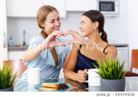 Young smiling emotional girlfriends showing the heart with hands and drinking tea Young smiling emotional girlfriends showing the heart with hands and drinking tea 59434926