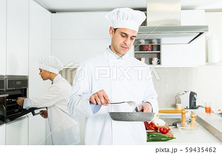 Male cook with stewpot working on kitchen with woman cook Male cook with stewpot working on kitchen with woman cook 59435462