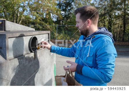 Young caucasian man in blue coat throwing away glass jar into a recycling container in Germany 59443925