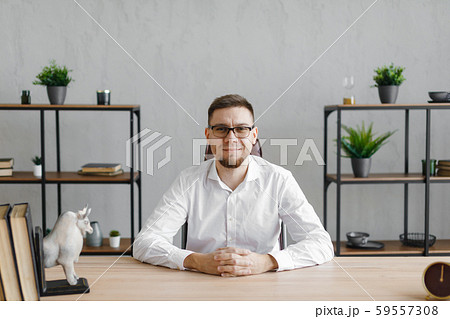 Portrait of young man sitting at his desk in the office and smiling at camera while sitting at Portrait of young man sitting at his desk in the office and smiling at camera while sitting at 59557308