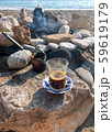 Transparent glass with coffee on a saucer. Coffee Turk and a steaming kettle in the background. Making coffee on a sandy beach among the stones. 59619179