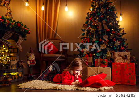 Little girl lying in carpet with presents around using tablet on red pillow. 59624285
