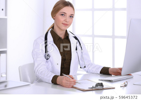 Young woman doctor at work in hospital looking at desktop pc monitor. Physician controls medication Young woman doctor at work in hospital looking at desktop pc monitor. Physician controls medication 59645372