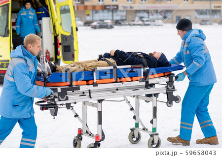 Two paramedics pushing sick unconscious man on stretcher towards ambulance car 59658043
