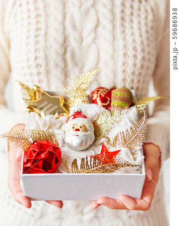 Woman in white knitted sweater is holding white box with New Year decorations. Bright red and golden decorative toys for Christmas tree. 59686938