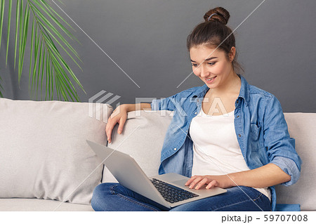 Freestyle. Young woman sitting on sofa studio isolated on grey browsing internet on laptop joyful 59707008