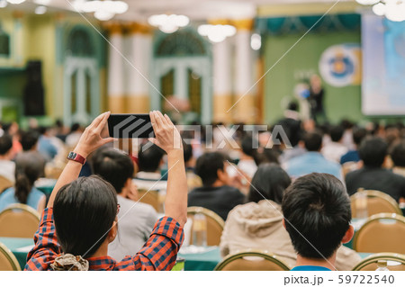 Closeup woman audience hand holding smart mobile phone for taking photo or doing Live streaming to Asian Speaker with casual suit on the stage present the screen in conference hall or seminar meeting 59722540