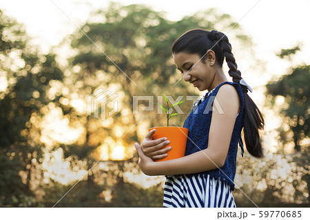 Low angle view of a smiling young girl holding a flower pot in hand standing in park and looking at it. 59770685
