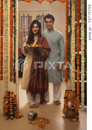 Happy young couple in traditional dress standing near the decorated entrance of their house with pooja plate. 59770708